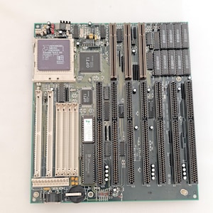 May include: A green and grey computer motherboard with various slots, chips, and connectors. The board features an Advanced Micro Devices processor and several Opti chips. It has numerous expansion slots and a battery.