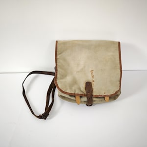 Peut inclure: Sac à bandoulière en toile beige avec bordure marron et une sangle assortie. Le sac a une fermeture à rabat avec une lanière en cuir et des chevilles en bois. Forme rectangulaire, idéal pour transporter des objets.