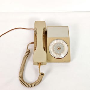 Puede incluir: Un teléfono de disco rotatorio beige vintage con auricular y cable en espiral. El teléfono tiene una base rectangular con un dial circular y una cuna separada para el auricular. Un diseño clásico de una época pasada.