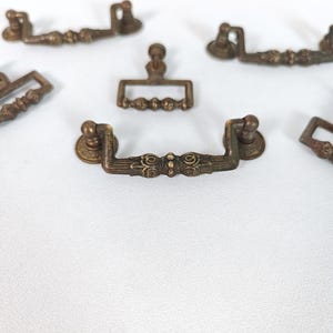 Op de afbeelding: Verschillende antieke bronzen lade-trekkers en een enkele trekker zijn gerangschikt op een wit oppervlak. De trekkers hebben een decoratief ontwerp met afgeronde uiteinden en sierlijke details. Geschikt voor meubelrestauratie.