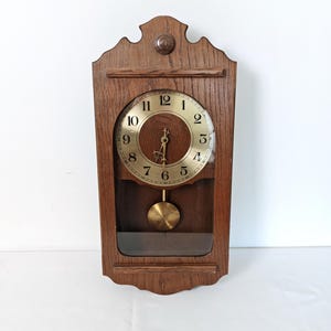 May include: A vintage wooden wall clock with a gold-colored clock face and black numerals. The clock has a pendulum and is encased in a dark brown wooden frame. The clock's design is traditional and features a decorative top.
