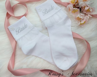 Puede incluir: Un par de calcetines blancos hasta el tobillo con un borde de encaje y la palabra "Bride" bordada en el puño. Los calcetines se muestran sobre una cinta rosa y una superficie blanca peluda.