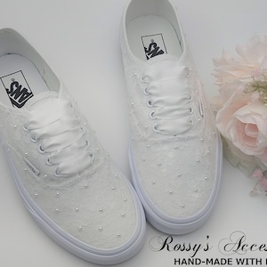 Könnte beinhalten: Ein Paar weiße Vans-Sneaker aus Spitze mit weißen Satinbändern und Perlenbesatz. Die Schuhe stehen auf einer weißen Oberfläche neben einem Strauß rosa Rosen.