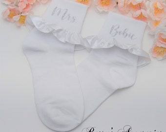 Puede incluir: Un par de calcetines blancos hasta el tobillo con un ribete de satén con volantes. Los calcetines están bordados con las palabras "Mrs" y "Babe" en purpurina plateada.