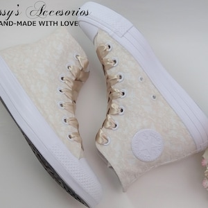 May include: A pair of white high-top sneakers with a cream-colored floral pattern and gold satin laces. The shoes are tied with a bow and the brand name "Converse" is visible on the side.