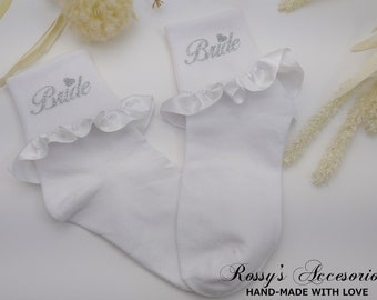 Puede incluir: Un par de calcetines blancos con un borde de volantes y la palabra "Bride" en purpurina plateada en la parte superior de cada calcetín.