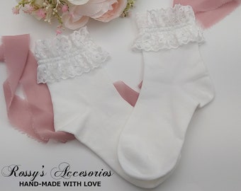 Puede incluir: Calcetines blancos hasta el tobillo con un borde de encaje blanco y detalles de perlas. Los calcetines están doblados y se muestran sobre un fondo de tela rosa. "Rossy's Accesorios HAND-MADE WITH LOVE" está escrito en texto negro en la parte inferior de la imagen.