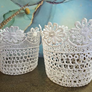 Peut inclure: Deux paniers en dentelle blanche faits à la main. Les paniers ont une forme cylindrique avec un motif ajouré délicat. Les bords sont ornés d'une garniture en dentelle florale. Le fond est un mélange de bleu et de vert.