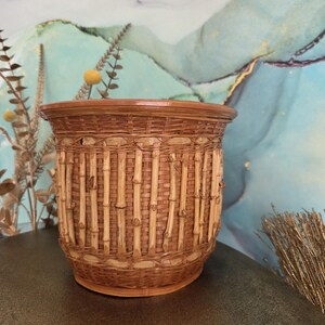Peut inclure: Un cache-pot cylindrique en osier tressé avec un extérieur en roseau brun clair et des tiges de bambou verticales disposées autour du centre. Le panier présente un bord large et renforcé ainsi qu'une base plate, dans des tons naturels de brun.