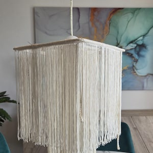 Peut inclure: Un lustre en macramé blanc, de forme carrée et à plusieurs niveaux. Le luminaire est suspendu au plafond et présente de longs brins de fil blanc cassé. Ce lustre est un élément décoratif pour la décoration intérieure.