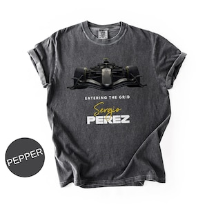 May include: Pepper-colored t-shirt featuring a Formula 1 race car graphic. The design includes the text "ENTERING THE GRID" above the name "Sergio PEREZ" in gold and white. The shirt has a vintage, worn look.