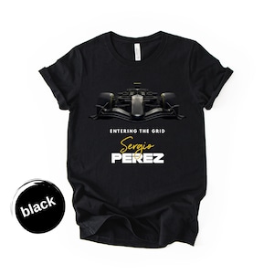 May include: Black t-shirt featuring a Formula 1 race car graphic. The shirt includes the text "ENTERING THE GRID" above the name "Sergio PEREZ" in white and gold lettering. A black circle with the word "black" is in the lower left corner.