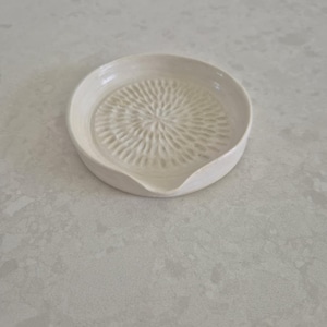 May include: A white ceramic garlic grater with a textured grating surface and a small spout. The round grater is designed for kitchen use and sits on a light-coloured surface.