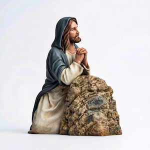Op de afbeelding: Een religieus beeldje dat een figuur met lang bruin haar en een baard afbeeldt, gekleed in een blauwe capuchon en witte kleding. De figuur knielt met gevouwen handen in gebed, leunend op een rots met textuur.
