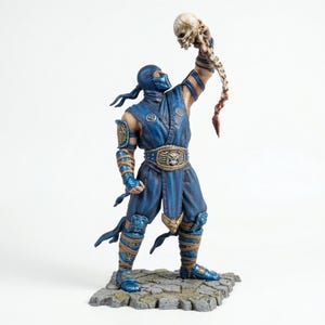 May include: A blue action figure of a masked warrior holding a skull aloft. The figure is clad in a blue outfit with gold accents and stands on a stone base. The skull is off-white with a bone-coloured spine.