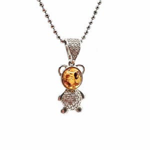 May include: A silver-toned necklace featuring a pendant in the shape of a teddy bear. The bear's body is encrusted with small, sparkling stones, and its belly is a round, amber-colored stone. The necklace chain is a delicate ball chain.