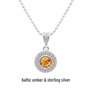 May include: A silver necklace with a round pendant featuring Baltic amber and sparkling stones. The pendant is encircled by a double row of small, clear stones. The necklace chain is a ball chain style. The text "baltic amber & sterling silver" is at the bottom.