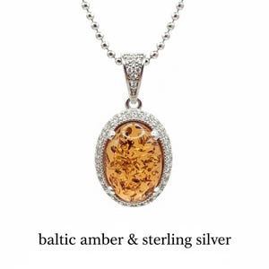 May include: A silver necklace with an oval pendant featuring Baltic amber. The amber is a warm, golden color with visible inclusions. The pendant is surrounded by a halo of small, clear stones and hangs from a delicate chain. The text "baltic amber & sterling silver" is below the pendant.