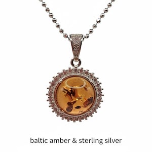 May include: A silver necklace with a round pendant featuring Baltic amber. The amber is a warm, golden color with inclusions. The pendant is surrounded by a halo of small, pink gemstones and the text "baltic amber & sterling silver".