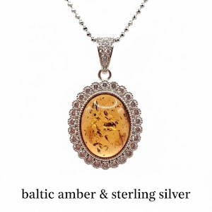 May include: A silver necklace with a pendant featuring an oval Baltic amber stone. The amber is encased in a decorative silver setting with a halo of small, pinkish-purple stones. The text "baltic amber & sterling silver" is at the bottom.