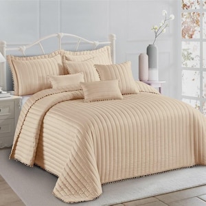May include: A beige bedspread and pillow set with a vertical ribbed design. The set includes a bedspread, several pillows, and a decorative trim. The bed is made up with a white metal frame and a matching bedside table with a lamp.
