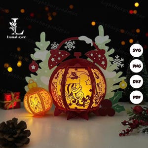 Può includere: Una lanterna di carta rossa con un disegno di Babbo Natale, illuminata dall'interno, è posta accanto a un ornamento rotondo più piccolo. Un ritaglio di albero di Natale e altre decorazioni festive sono sullo sfondo. Il testo "LumaLayer" è incluso.