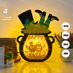 Puede incluir: Una pieza de arte en papel en capas que representa una olla de oro negra con un sombrero y piernas de duende. La olla tiene las palabras "HAPPY" y "GO LUCKY". La obra de arte está iluminada con un brillo cálido.