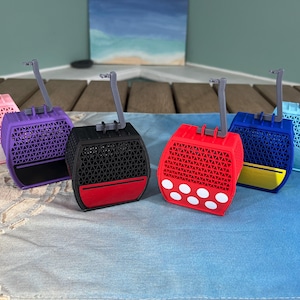 May include: A collection of colorful, miniature cable car models. The models are in various colors, including pink, purple, black, red, blue, and light blue. Each has a detailed geometric pattern and a small, gray support arm. The cable cars are displayed on a blue surface.