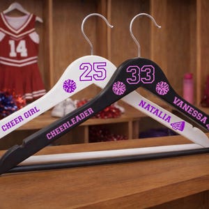 Peut inclure: Deux cintres en bois, un blanc et un noir, personnalisés pour les cheerleaders. Le cintre blanc porte le numéro 25 et les mots "CHEER GIRL". Le cintre noir porte le numéro 33 et les noms "NATALIA" et "VANESSA".