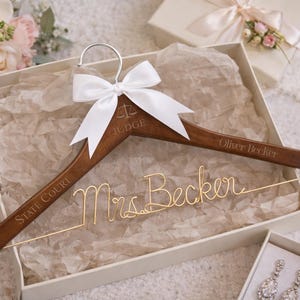 May include: A wooden clothes hanger with the words "State Court" and "Judge" engraved. A white bow is tied at the top. The name "Oliver Becker" is engraved on the right. "Mrs. Becker" is written in gold wire.