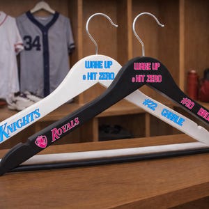May include: Two wooden clothes hangers, one black and one white, with silver hooks. The white hanger has "Knights" and "Wake Up & Hit Zero" printed on it. The black hanger has "Royals", "Wake Up & Hit Zero", "#22 Charlie", and "#10 Brock" printed on it.
