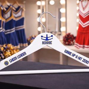 Peut inclure: Un cintre en bois blanc avec le nom "ROHHIE" et les mots "CHEER LEADER" et "WAKE UP & HIT ZERO" en bleu. Le cintre a un crochet argenté et un emblème bleu et jaune. Des uniformes de pom-pom girl sont visibles en arrière-plan.