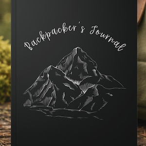 May include: Black journal featuring the words "Backpacker's Journal" in cursive. A white mountain range illustration is below the text. The journal is set against a blurred background.