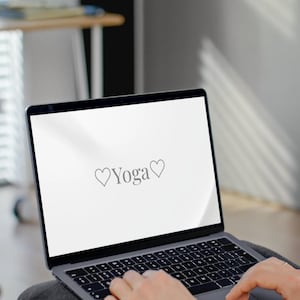May include: A silver laptop displaying the word "Yoga" with a heart symbol on a white screen. The laptop is open, and the keyboard is visible. The image is taken indoors, with a blurred background.