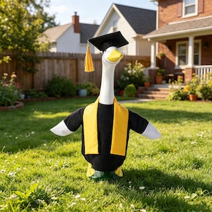 May include: A goose statue dressed in graduation attire, including a black cap with a gold tassel and a yellow sash. The goose has a white neck and yellow beak, standing on green grass.