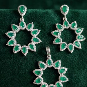 May include: A jewelry set consisting of two drop earrings and a matching pendant. Each piece features a circular arrangement of pear-shaped green stones surrounded by small clear stones, set in a silver-toned metal frame with a polished finish.