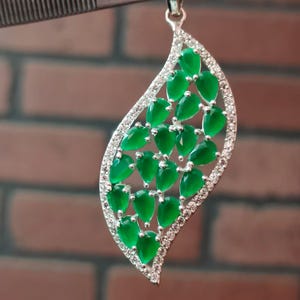 May include: A leaf-shaped pendant featuring multiple pear-cut green gemstones arranged in a cluster. The outer edge of the pendant is lined with small, clear, sparkling stones set in a silver-toned metal frame with a bail at the top.