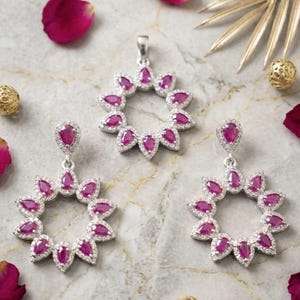 May include: A jewelry set consisting of a pendant and two matching drop earrings. Each piece features a circular arrangement of pear-shaped, deep pink gemstones surrounded by small, clear, sparkling stones set in a silver-toned metal frame.