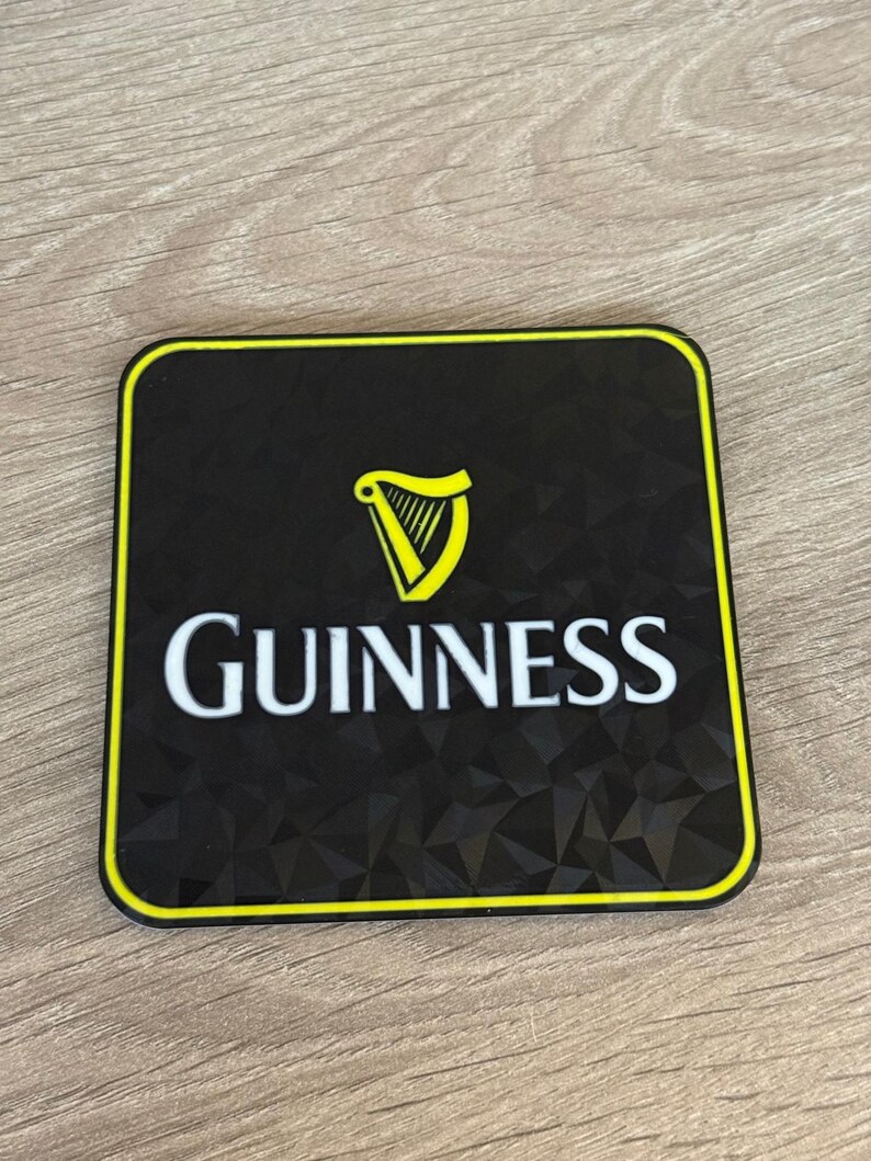 以下が含まれることがあります： 黒い正方形のコースターで、黄色の縁取りがあります。「Guinness」の文字が白で、その上に黄色のハープのロゴがあります。コースターの表面はテクスチャ加工されています。