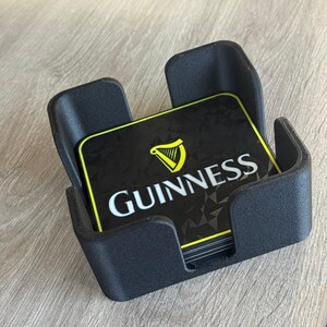 以下が含まれることがあります： 黒いコースターホルダーとギネスコースターのセット。 コースターは黒で、黄色の縁取りと白いギネスのロゴが付いています。 ホルダーは暗い素材でできており、明るい木製の表面に置かれています。