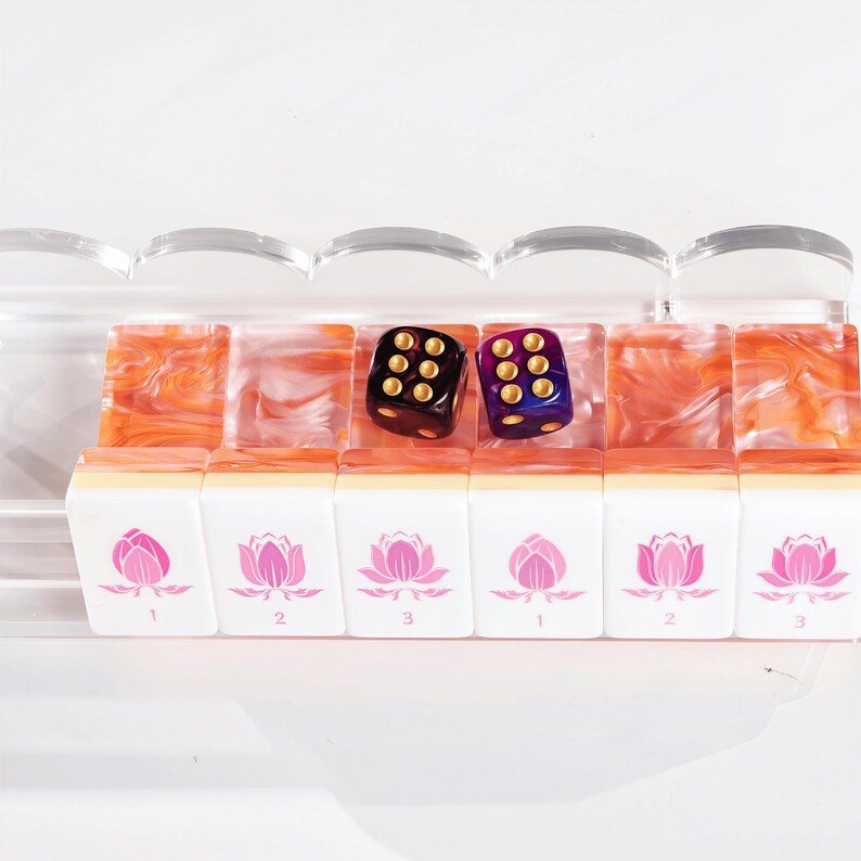 Puede incluir: Un conjunto de seis fichas de mahjong rectangulares con caras blancas que presentan ilustraciones de flores de loto rosas y n&uacute;meros, dispuestas en un soporte de acr&iacute;lico transparente. Dos dados de seis caras, uno negro y otro morado con puntos dorados, descansan sobre las fichas.