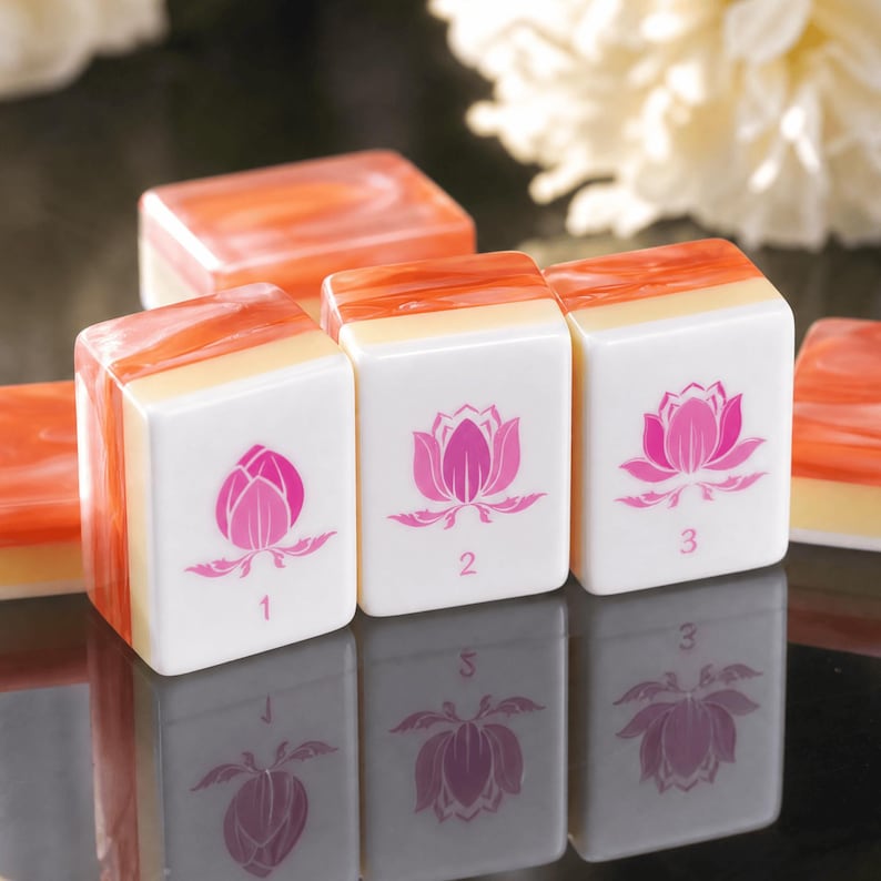 Puede incluir: Tres fichas de mahjong rectangulares con caras blancas y partes superiores de color naranja jaspeado. Cada ficha presenta un gr&aacute;fico de flor de loto rosa y un n&uacute;mero: 1, 2 y 3. Las fichas est&aacute;n dispuestas en fila sobre una superficie oscura.