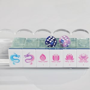 May include: A set of six white mahjong tiles arranged in a clear acrylic rack. The tiles feature blue and purple dragon illustrations and pink lotus flower designs with the numbers 1 through 4. Two six-sided dice, one blue and one purple, rest on top.