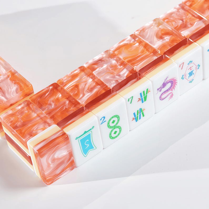 Puede incluir: Un juego de fichas de mahjong con el reverso de acr&iacute;lico transl&uacute;cido veteado en naranja y blanco y el frente blanco. Las fichas muestran varios s&iacute;mbolos grabados en color, incluyendo una bandera azul, un c&iacute;rculo verde, tallos de bamb&uacute;, un drag&oacute;n morado y caracteres chinos.