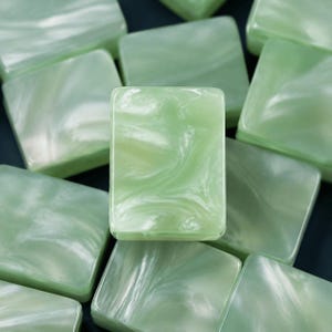 May include: A collection of rectangular buttons featuring a smooth, polished surface with a marbled pattern in shades of pale mint green and white. Each button has a glossy finish and a consistent, uniform shape, arranged in a layered, overlapping stack.