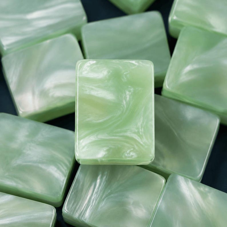 May include: A collection of rectangular buttons featuring a smooth, polished surface with a marbled pattern in shades of pale mint green and white. Each button has a glossy finish and a consistent, uniform shape, arranged in a layered, overlapping stack.