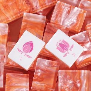 Puede incluir: Una colecci&oacute;n de fichas de mahjong rectangulares con el reverso de resina veteada en naranja y blanco. Dos fichas se sit&uacute;an en primer plano, mostrando una cara blanca con un gr&aacute;fico de flor de loto rosa y los n&uacute;meros 1 y 2.