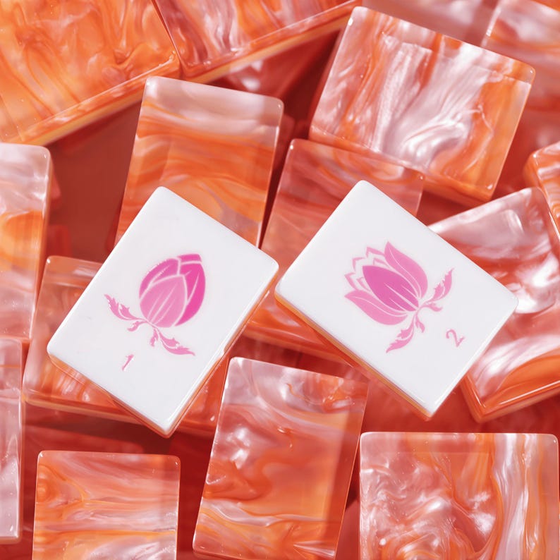 Puede incluir: Una colecci&oacute;n de fichas de mahjong rectangulares con el reverso de resina veteada en naranja y blanco. Dos fichas se sit&uacute;an en primer plano, mostrando una cara blanca con un gr&aacute;fico de flor de loto rosa y los n&uacute;meros 1 y 2.