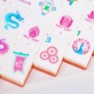 Puede incluir: Una colecci&oacute;n de fichas de mahjong rectangulares blancas con coloridos dise&ntilde;os impresos que incluyen un drag&oacute;n azul, flores de loto rosas, un pavo real, un estandarte con la letra e y tres monedas circulares con centros cuadrados, todas con bases naranjas.