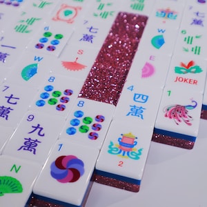 May include: A collection of white Mahjong tiles featuring colorful printed designs including Chinese characters, numbers, fans, a peacock, and a joker mask. The tiles have a dark blue base and a strip of pink glitter embedded along the side.